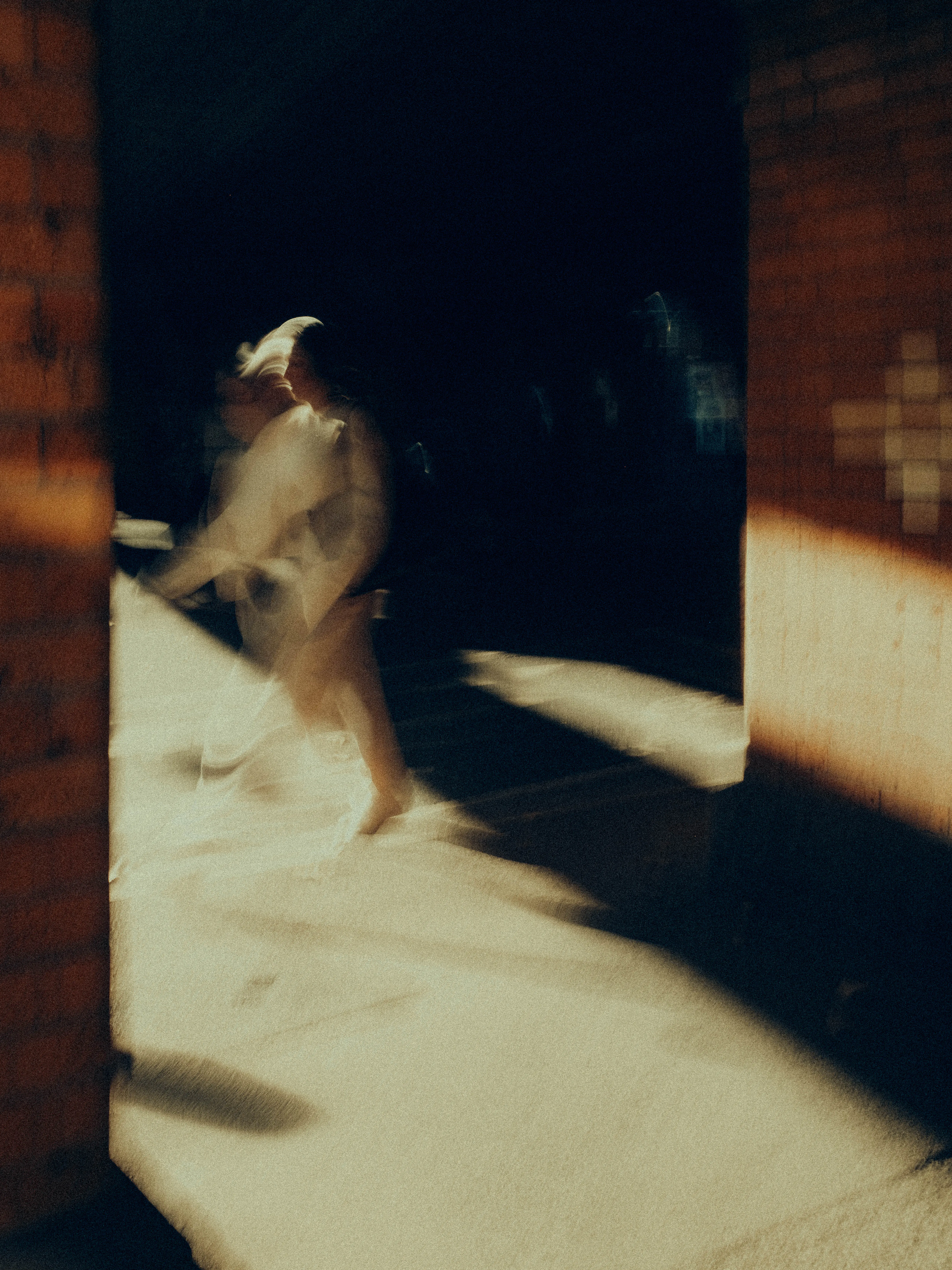A figure in pale clothing dissolves through a shaft of harsh golden sunlight framed by dark brick walls, their movement becoming pure luminous energy against the deep surrounding shadow.
