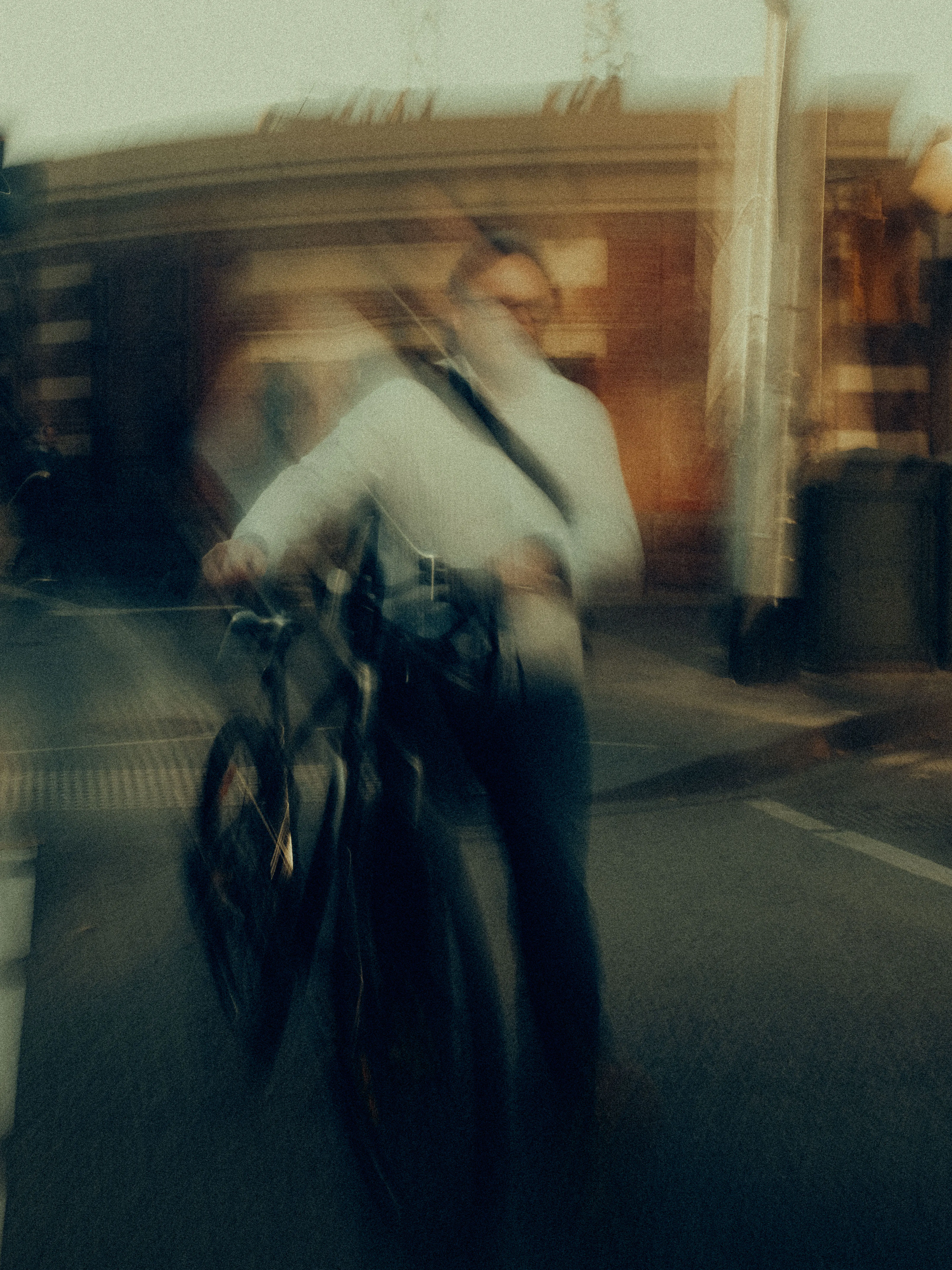 A cyclist pauses mid-motion on a city street at dusk, body and bicycle rendered as warm layered blur against the terracotta brick of a corner building in fading golden light.
