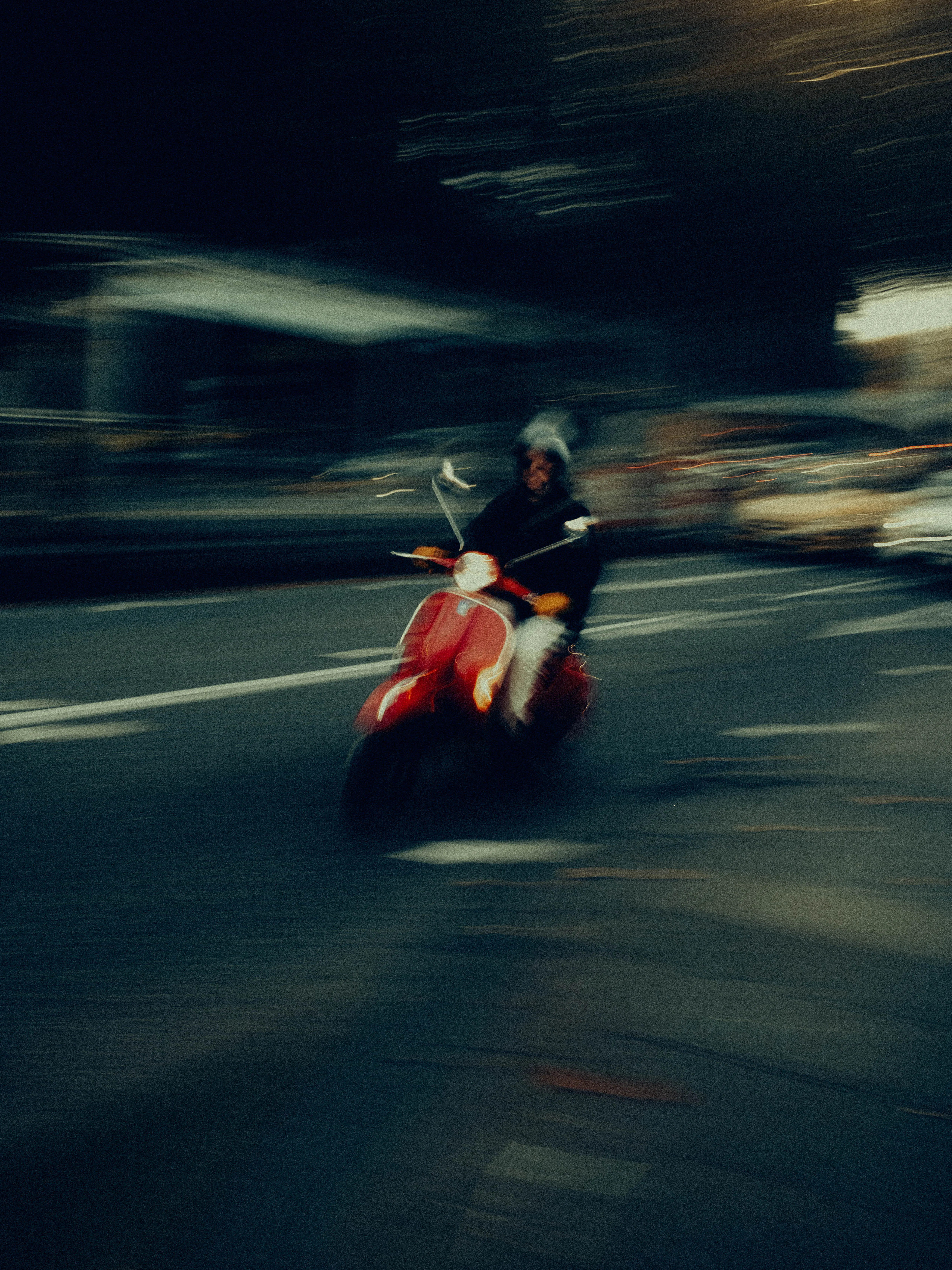 A red scooter blazes through a dark city street at night, its rider reduced to a smear of motion against streaked headlights and deep shadow, the scene almost entirely consumed by speed.