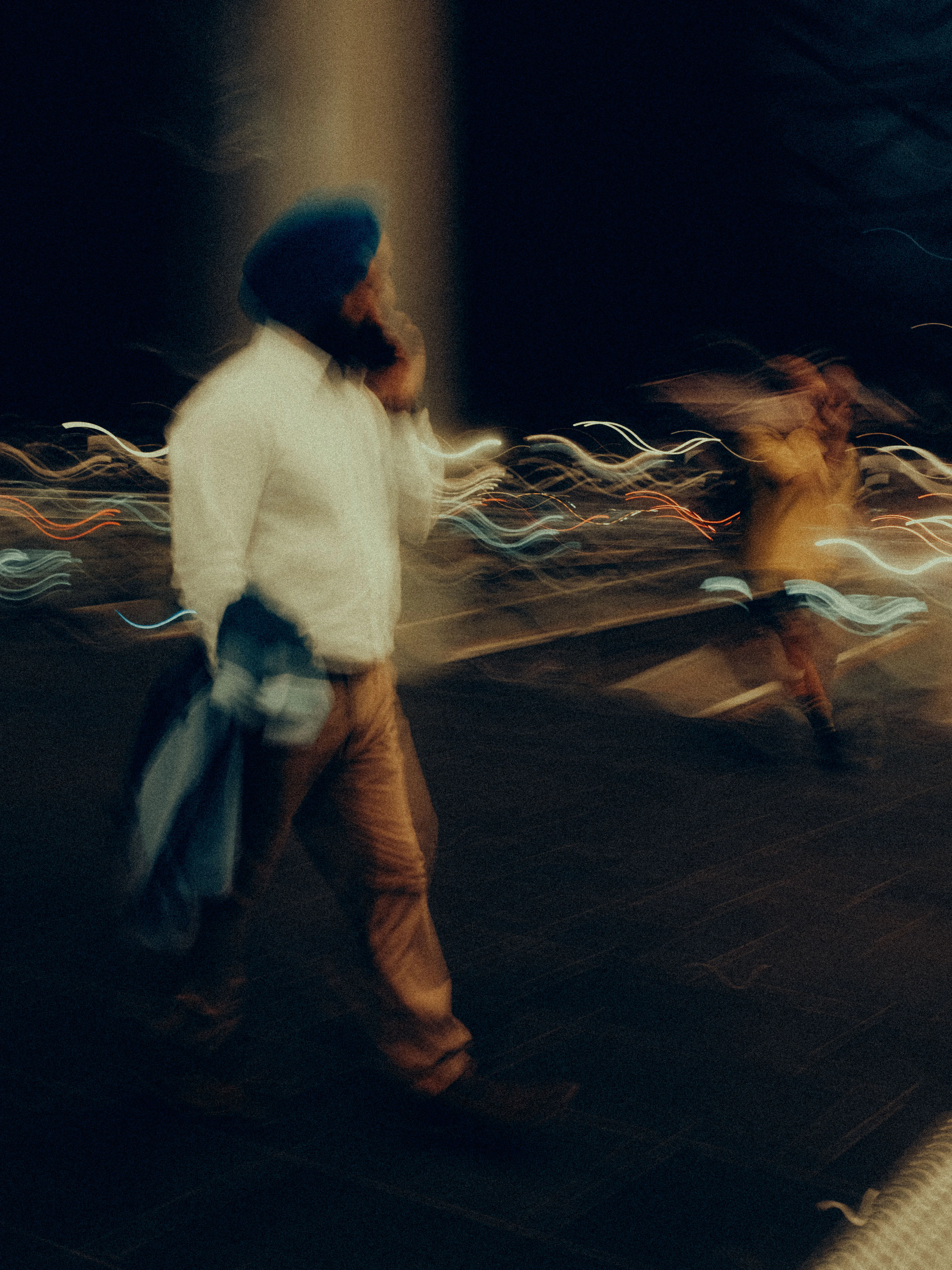 Two pedestrians — one in a blue turban and white jacket, another in amber — stride through a night street as orange and teal light trails weave sinuously around their blurred forms.
