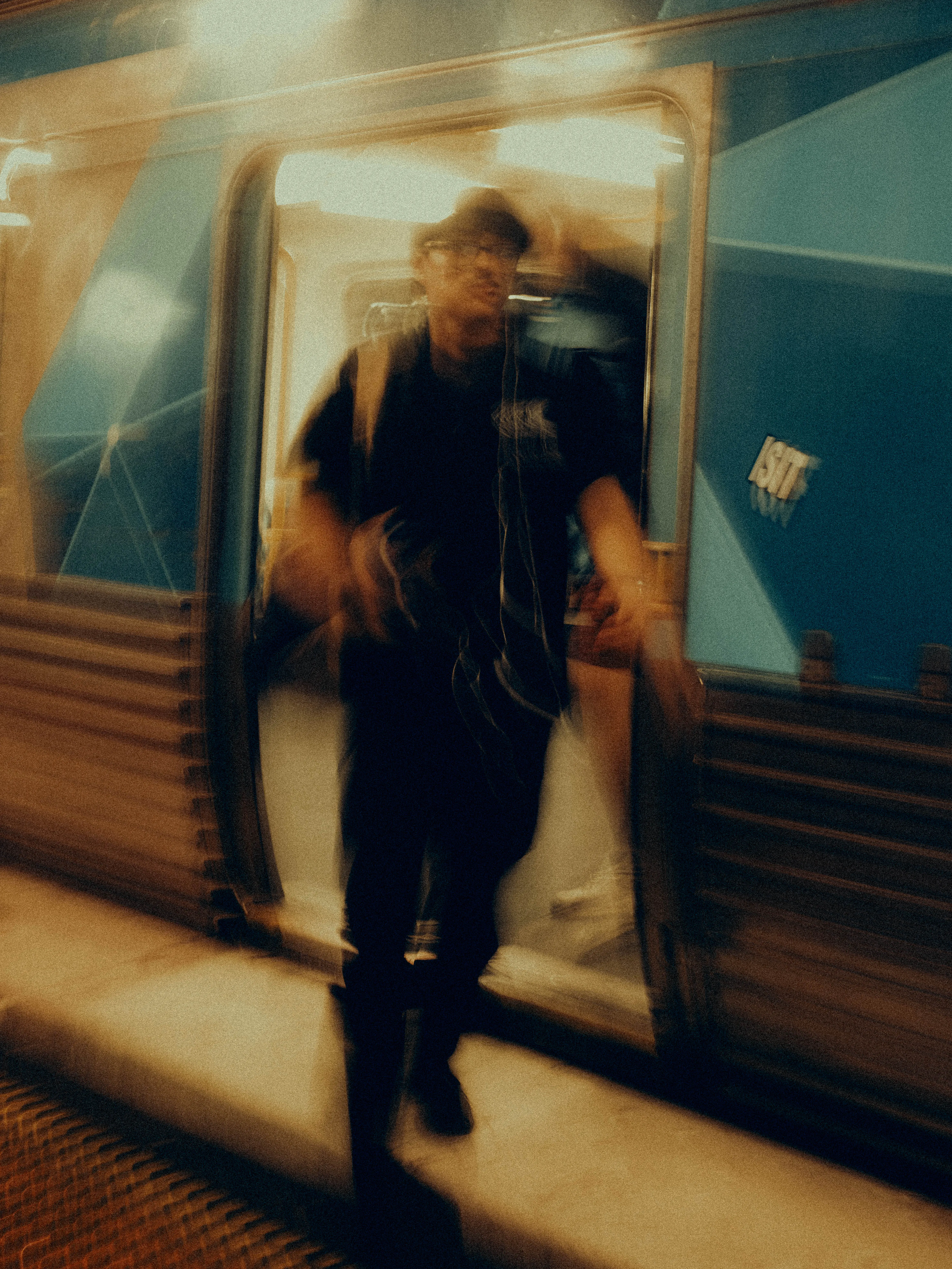 A hatted figure steps from the open door of a blue Melbourne tram onto a warmly lit platform, their form ghosted and doubled by the slow shutter into something between arrival and departure.