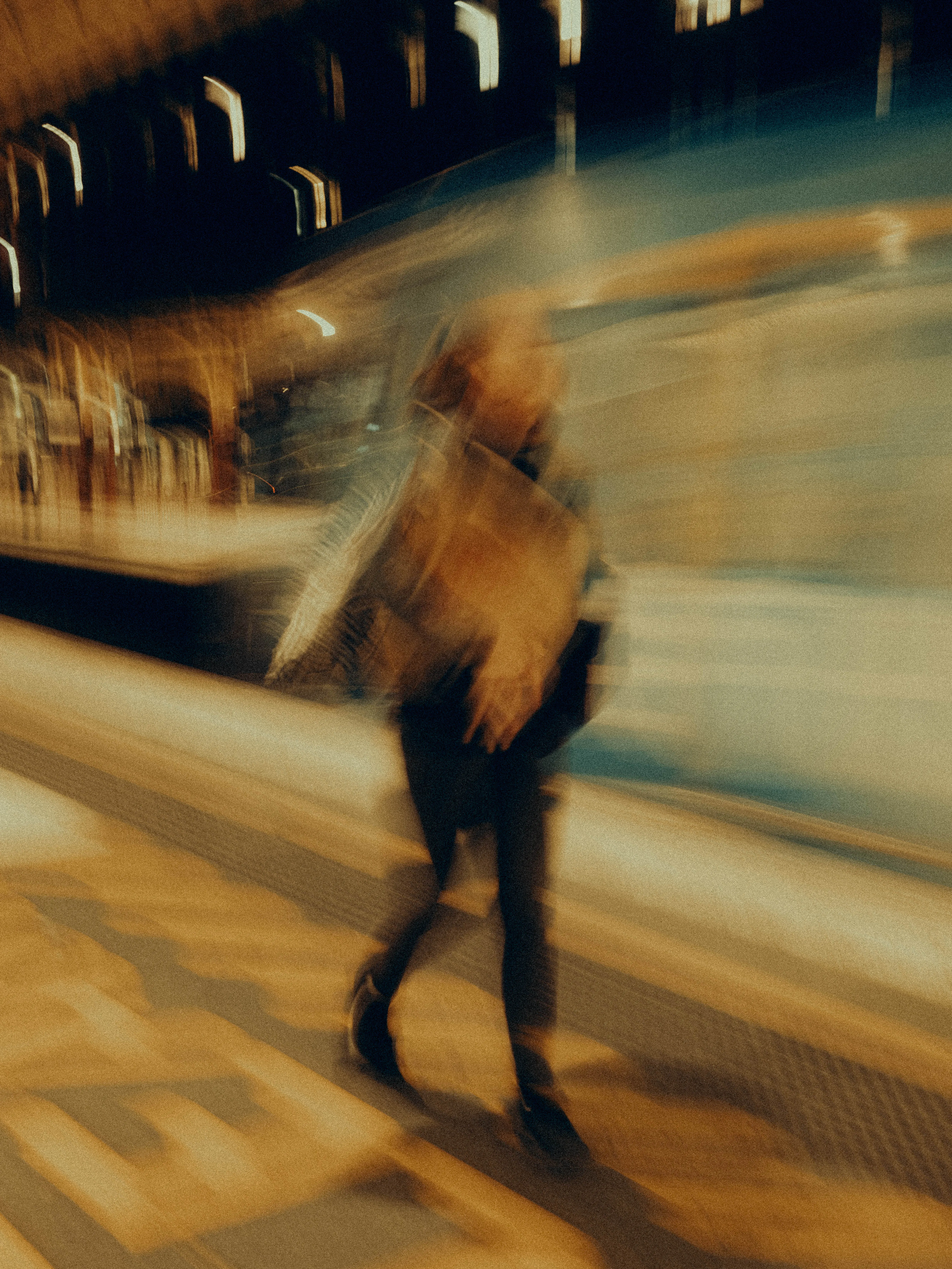 A lone figure carrying a bag strides along a golden train platform, the warm amber architecture sweeping diagonally behind them as motion blur dissolves the scene into kinetic abstraction.