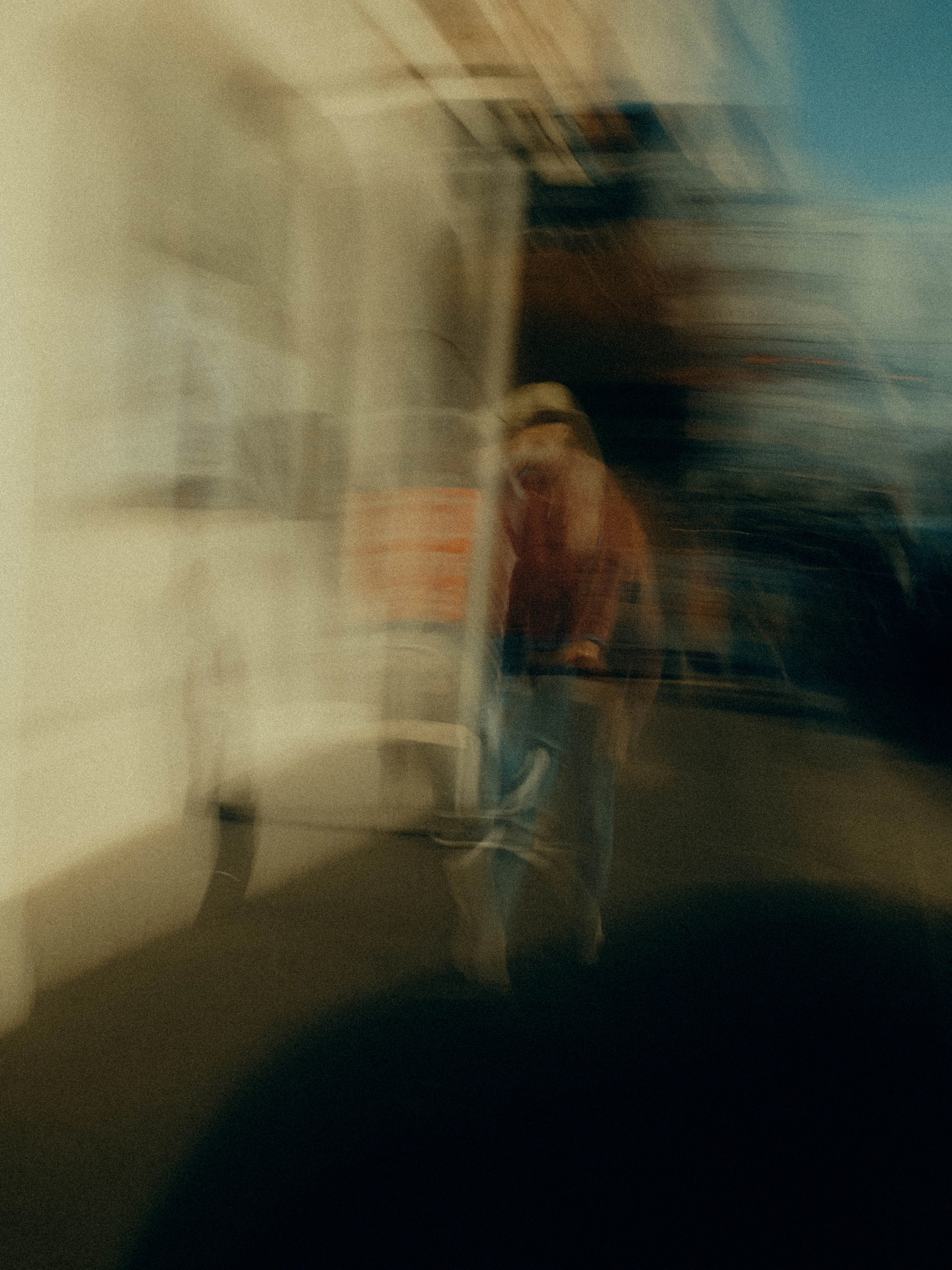 A figure in a wide-brimmed hat and red jacket moves through bright midday blur, pale light washing the architecture into cascading streaks of cream and open sky blue.