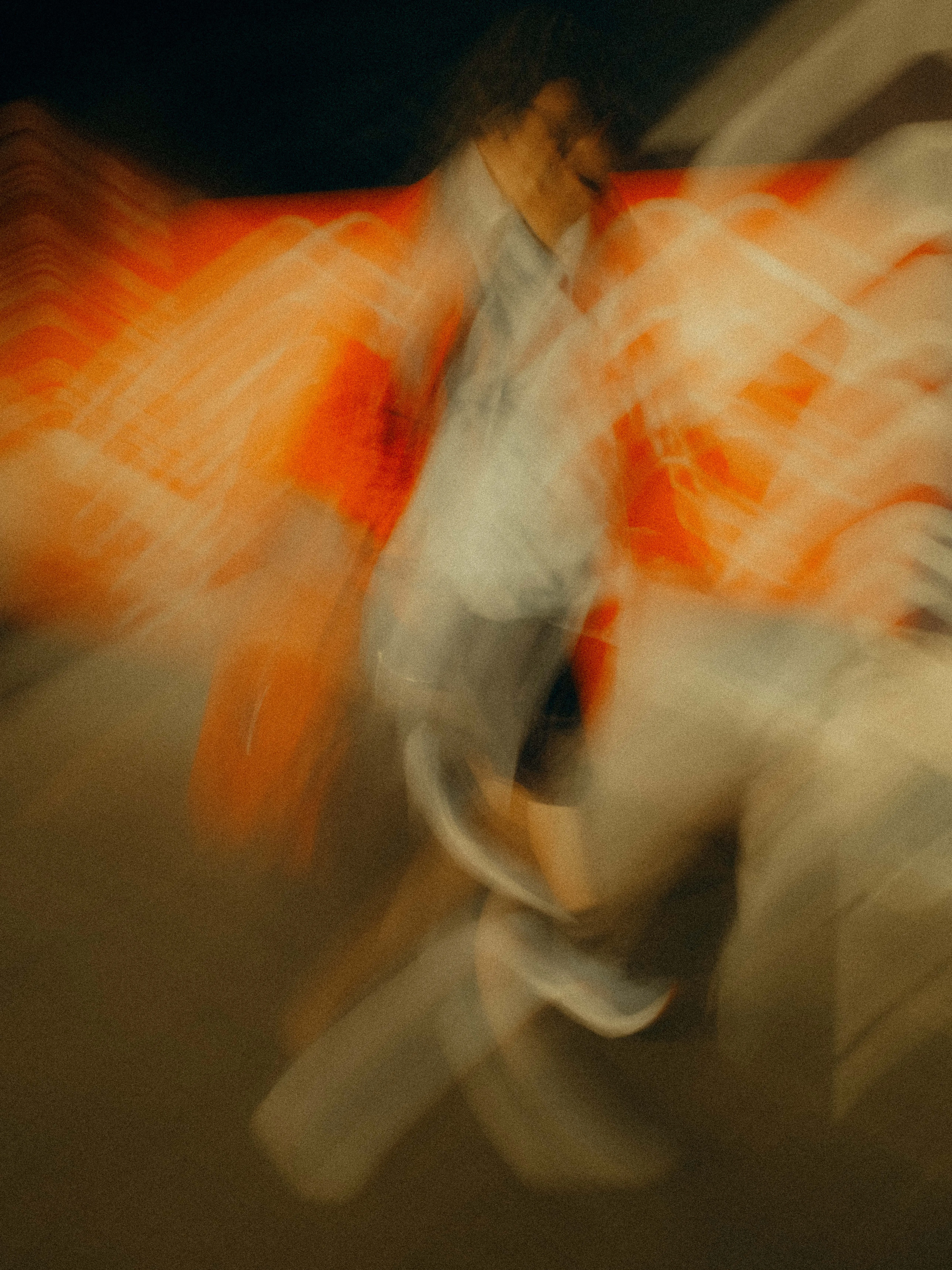 Two figures blur together in a swirl of vivid orange neon and white fabric, their forms barely separable from the city signage burning hot behind them in deep saturated abstraction.