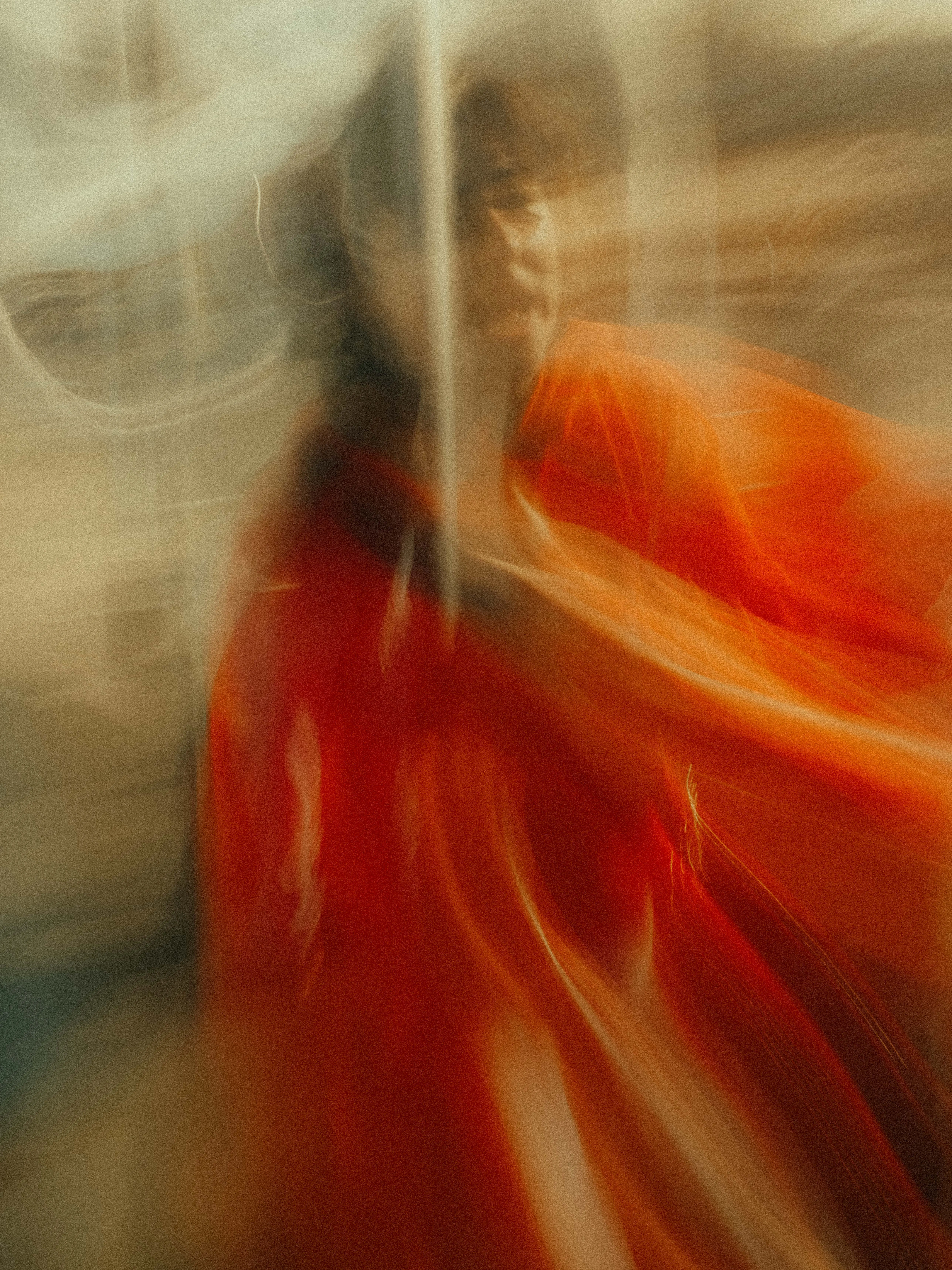 A figure swathed in billowing orange-red fabric dissolves into cascading streaks of flame and cream, their face barely legible through layered planes of intentional motion against a pale Melbourne street.