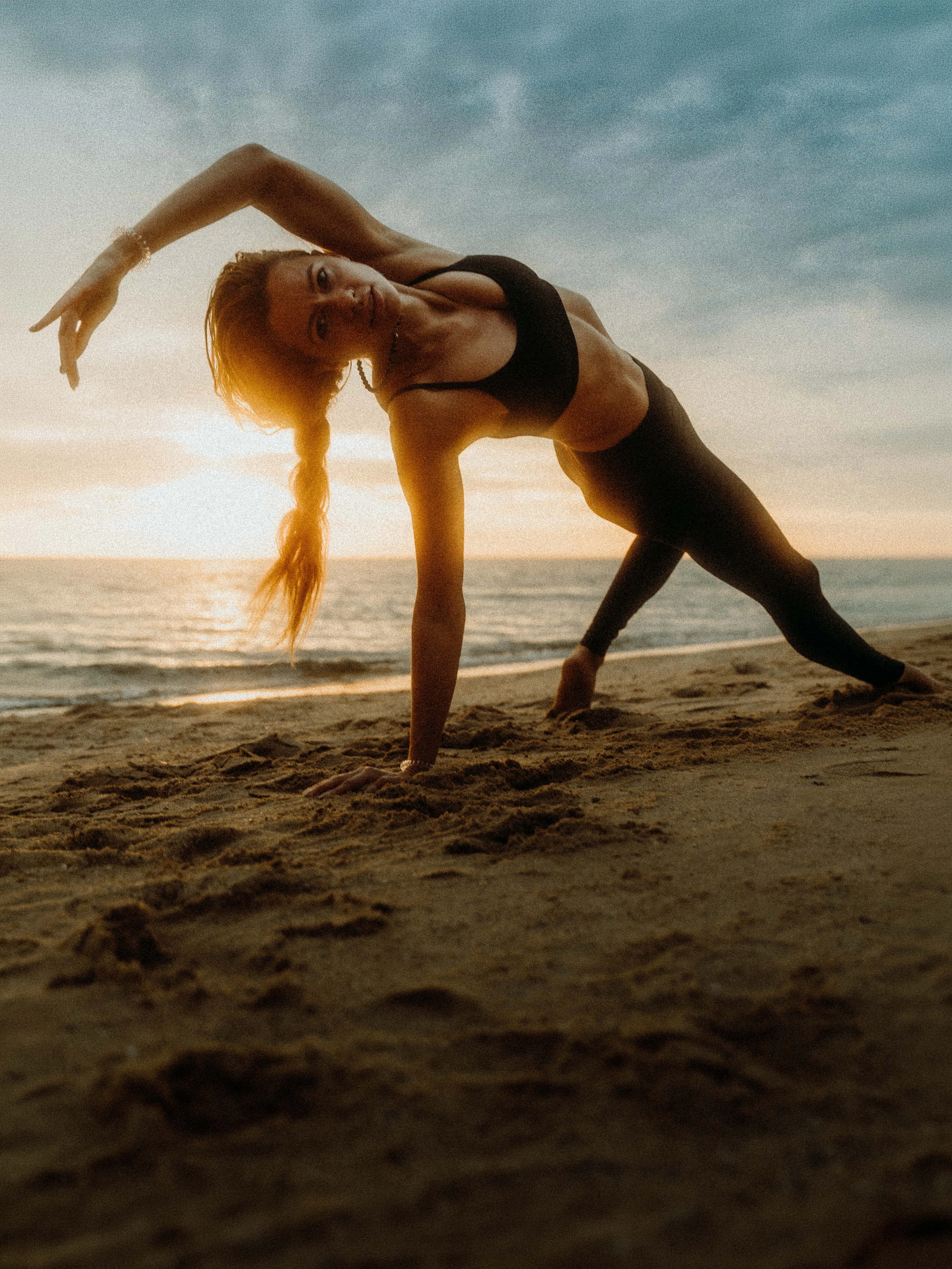 A woman in a black sports top and leggings holds a deep backbending yoga pose on a beach at sunrise, one arm arced overhead and hair falling loose, rim-lit gold by the low sun over a calm sea.